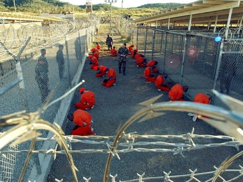 Detainees in orange jumpsuits sit in a holding area under the watch of U.S. military police at the temporary Camp X-Ray, which was later closed and replaced by Camp Delta, inside Guantanamo Bay naval base.(Handout/Reuters)