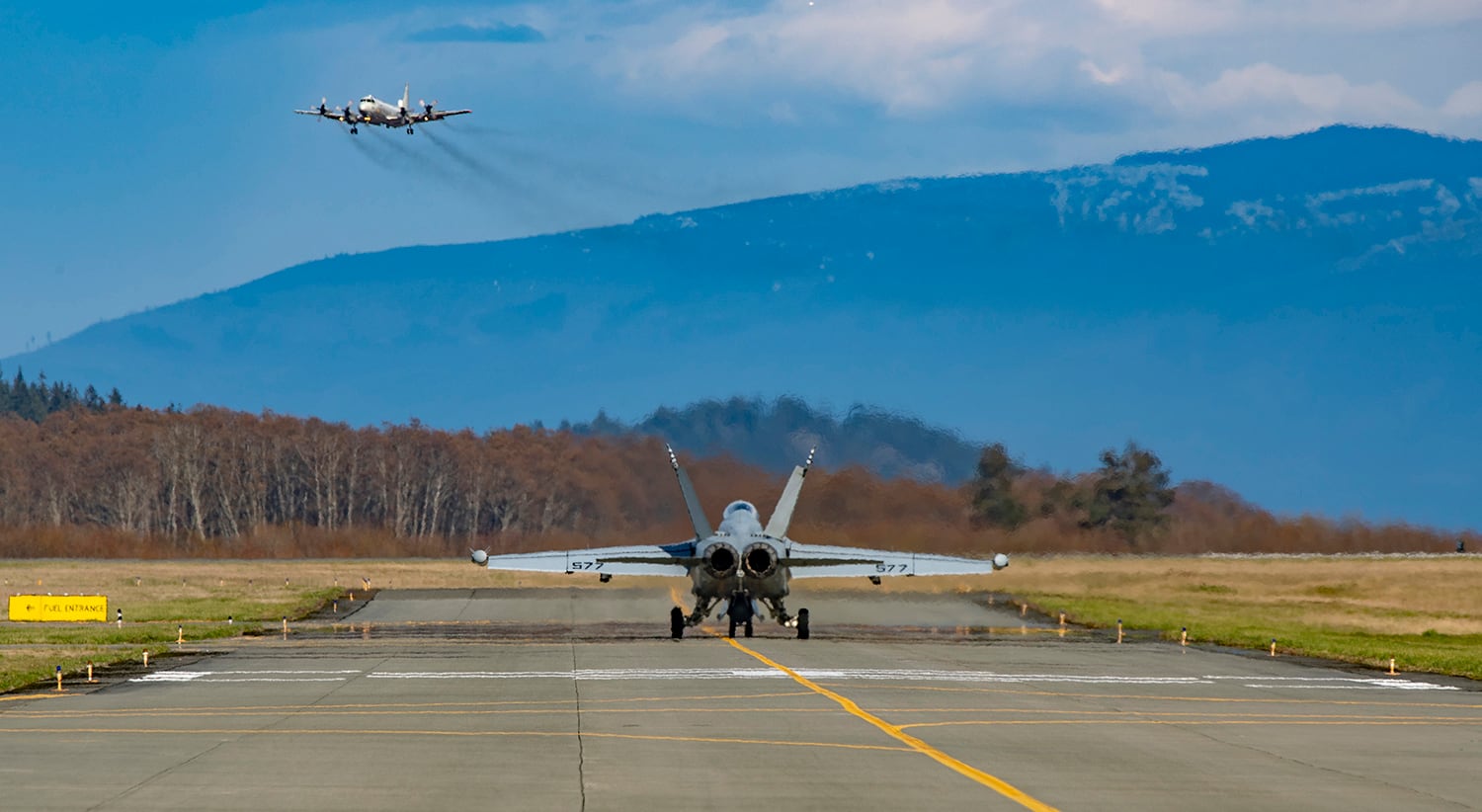 An EA-18G Growler taxis toward the runway on March 29, 2019, at Ault Field at Whidbey Island, Wash., as a P3C Orion performs a low approach.